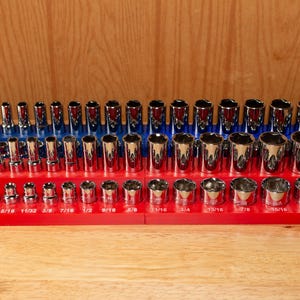 May include: A set of chrome sockets in various sizes, arranged on red and blue holders. The sockets are displayed on a wooden surface, showcasing a range of sizes for mechanical work. The image highlights the tools' shiny, reflective surfaces.
