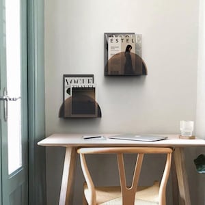 May include: Two black metal magazine holders with a half-circle design are mounted on a white wall. The holders are filled with magazines, including Vogue Travel and Estel.