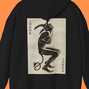 May include: Black hoodie with a white graphic of a joker card featuring a black and white illustration of a jester with a tail and the word "JOKER" printed vertically on either side of the illustration.