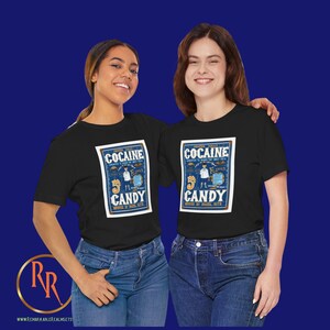 May include: Two people wearing black t-shirts with a vintage-style graphic design featuring the text "Cocaine Candy" and an illustration of a man in a lab coat. The graphic is surrounded by a blue border and has a gold background.