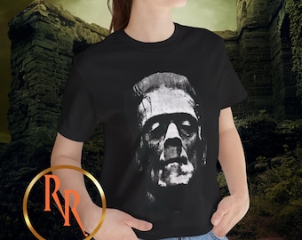 May include: Black t-shirt featuring a grayscale image of Frankenstein's monster. The graphic is a detailed portrait of the iconic character. The shirt is worn by a person in blue jeans. The website address is visible at the bottom of the image.