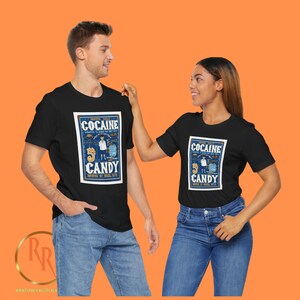 May include: Two people wearing black t-shirts with a vintage-style graphic design featuring the text "Cocaine Candy" and an illustration of a person in a blue suit holding a bag of candy. The graphic is set against a blue background with white and orange accents.