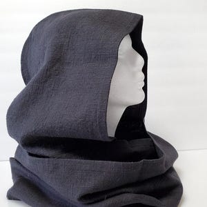 May include: A dark gray hooded cowl made of textured fabric. The cowl is draped over a white mannequin head, showcasing the hood's shape and the neck warmer design. The fabric appears to be linen or a similar material.