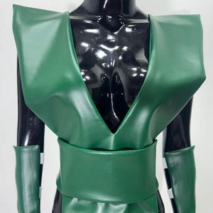 Puede incluir: Un disfraz verde con una parte superior con cuello en V, cinturón ancho y mangas a juego. El disfraz se exhibe en un maniquí negro. Las mangas tienen rayas blancas y negras.