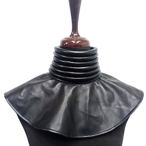 May include: A black faux leather neck accessory featuring a high, multi-layered ribbed collar that transitions into a wide, smooth, circular cape-like yoke that rests over the shoulders. The piece has a structured, layered design.
