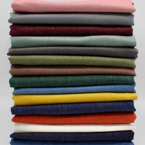 May include: A stack of folded fabric in various colors, including pink, gray, burgundy, green, yellow, and navy blue. The fabrics are neatly arranged, showcasing a range of hues suitable for sewing or crafting projects. The image is well-lit, highlighting the textures and colors.