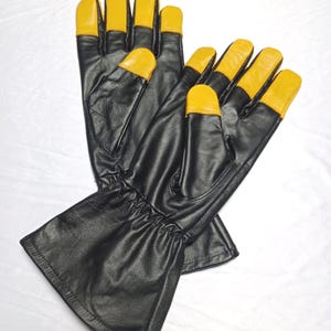 May include: A pair of black leather gloves with yellow fingertips. The gloves have a gathered wrist and are set against a white background. The gloves appear to be new.