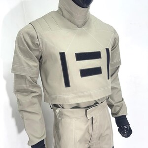 Puede incluir: Un atuendo futurista de color beige, que consta de una chaqueta corta y pantalones a juego. La chaqueta presenta detalles rectangulares negros y costuras blancas. El atuendo se exhibe en un maniqu&iacute; con cabeza y manos negras.