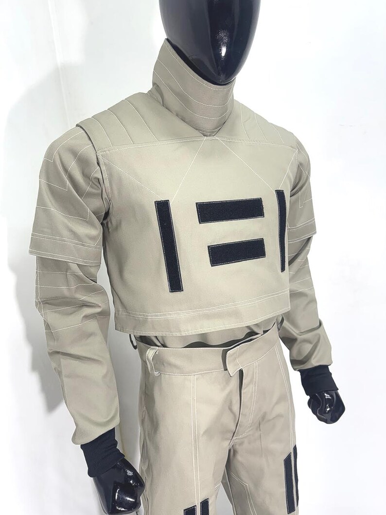 Puede incluir: Un atuendo futurista de color beige, que consta de una chaqueta corta y pantalones a juego. La chaqueta presenta detalles rectangulares negros y costuras blancas. El atuendo se exhibe en un maniqu&iacute; con cabeza y manos negras.