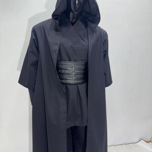 Op de afbeelding: Een zwart kostuum met capuchon en brede riem. Het kostuum bevat een lange, vloeiende jurk met capuchon, een bijpassende tuniek, broek en een brede, zwarte riem met zilverkleurige klinknagels. Het kostuum wordt op een mannequin getoond.