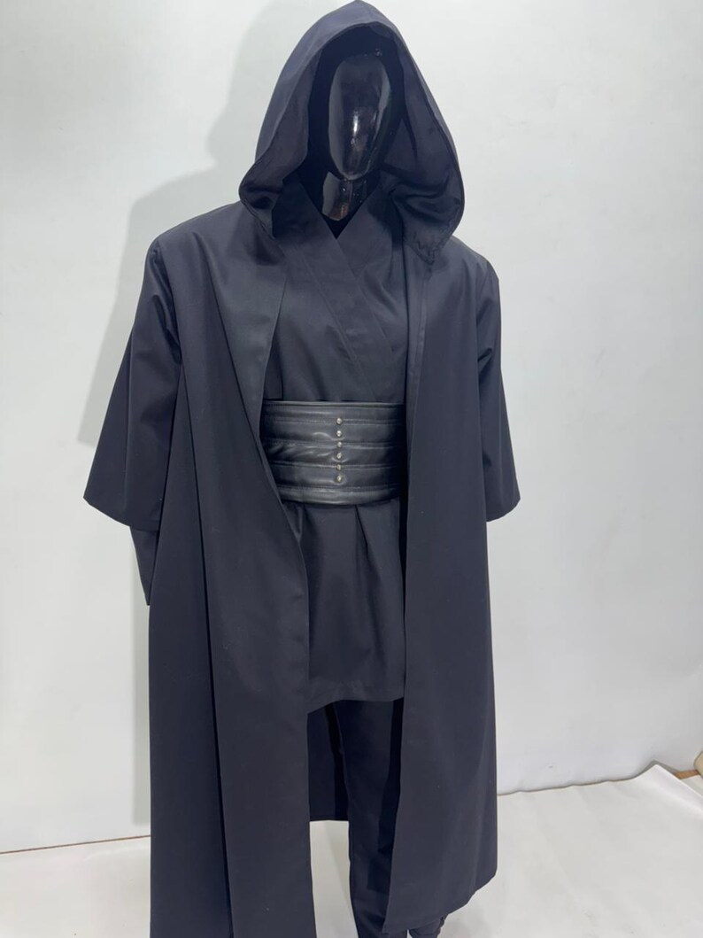 Op de afbeelding: Een zwart kostuum met capuchon en brede riem. Het kostuum bevat een lange, vloeiende jurk met capuchon, een bijpassende tuniek, broek en een brede, zwarte riem met zilverkleurige klinknagels. Het kostuum wordt op een mannequin getoond.