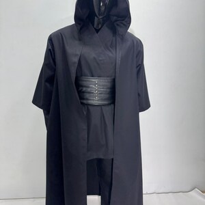 Op de afbeelding: Een zwart kostuum met bijpassende capuchonjas. Het kostuum bevat een lange, vloeiende jas met een grote capuchon, een tuniek, een broek en een brede, zwarte riem. De outfit wordt gepresenteerd op een zwarte mannequin.
