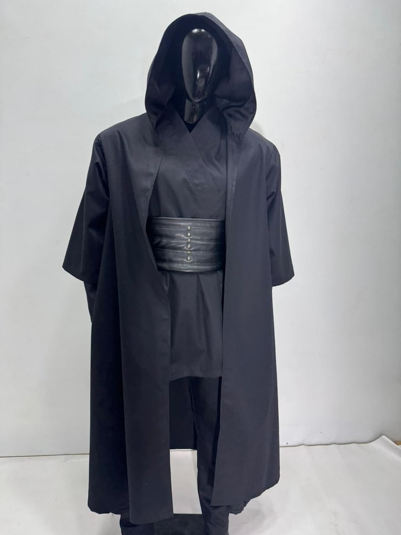 Op de afbeelding: Een zwart kostuum met bijpassende capuchonjas. Het kostuum bevat een lange, vloeiende jas met een grote capuchon, een tuniek, een broek en een brede, zwarte riem. De outfit wordt gepresenteerd op een zwarte mannequin.