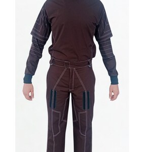 May include: A brown two-piece outfit featuring a high-neck top and matching pants. The top has short sleeves over long sleeves, and the pants have pockets. White stitching outlines the seams and details of the outfit. The outfit is displayed against a white background.