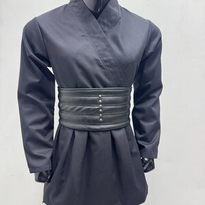 Op de afbeelding: Zwart vechtsportuniform op een mannequin. De outfit bestaat uit een wikkel-top, lange mouwen en een bijpassende broek. Een brede, zwarte riem met decoratieve klinknagels accentueert de taille. De stof heeft een lichte glans.