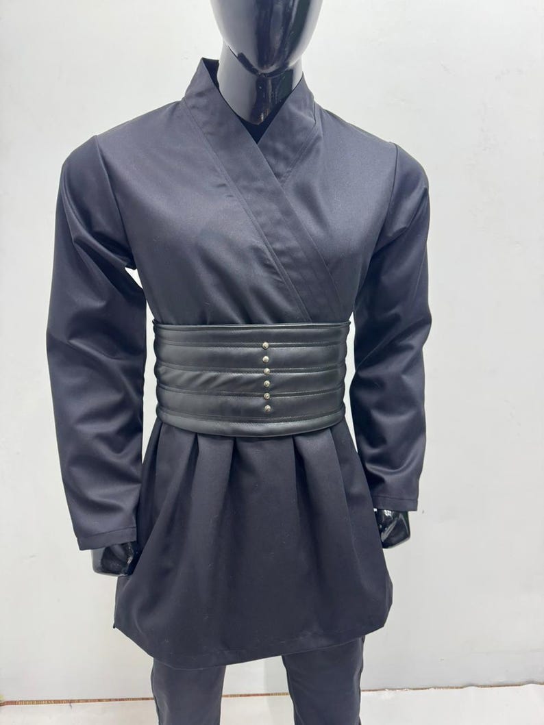 Op de afbeelding: Zwart vechtsportuniform op een mannequin. De outfit bestaat uit een wikkel-top, lange mouwen en een bijpassende broek. Een brede, zwarte riem met decoratieve klinknagels accentueert de taille. De stof heeft een lichte glans.
