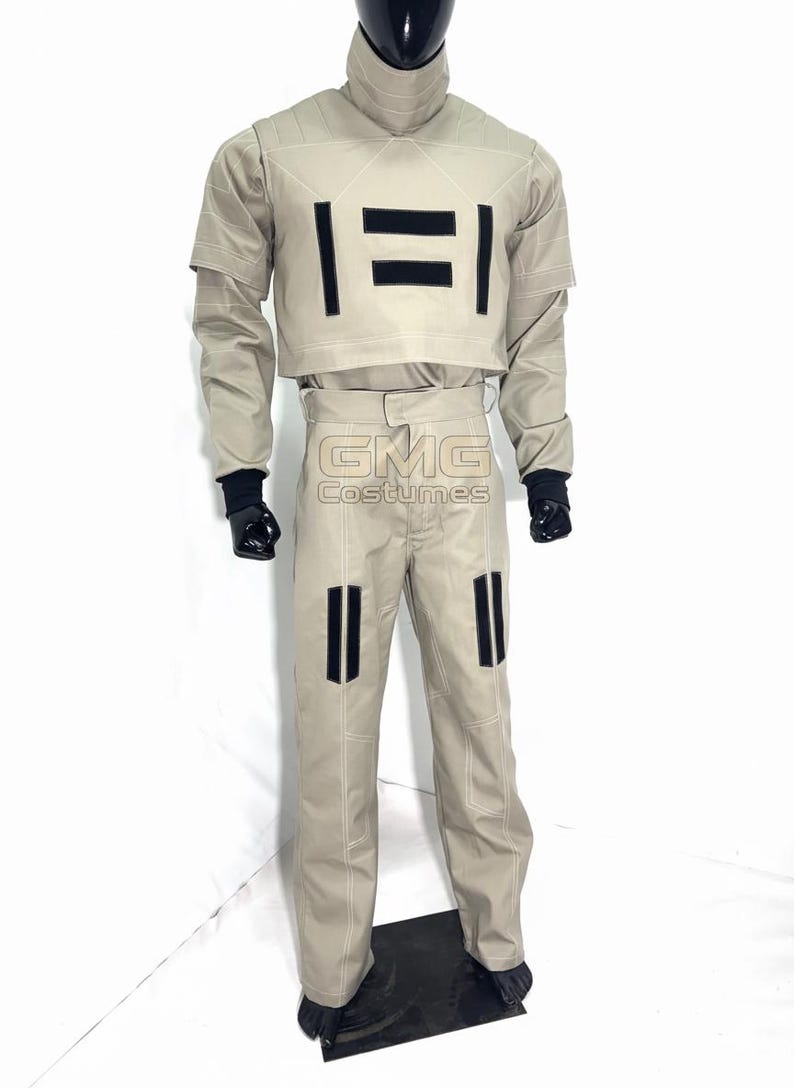 Puede incluir: Un disfraz de color beige con detalles en negro, que incluye una chaqueta corta y pantalones a juego. La chaqueta tiene dos rayas negras horizontales, y los pantalones tienen dos rayas verticales. El disfraz tiene el texto "GMG Costumes".