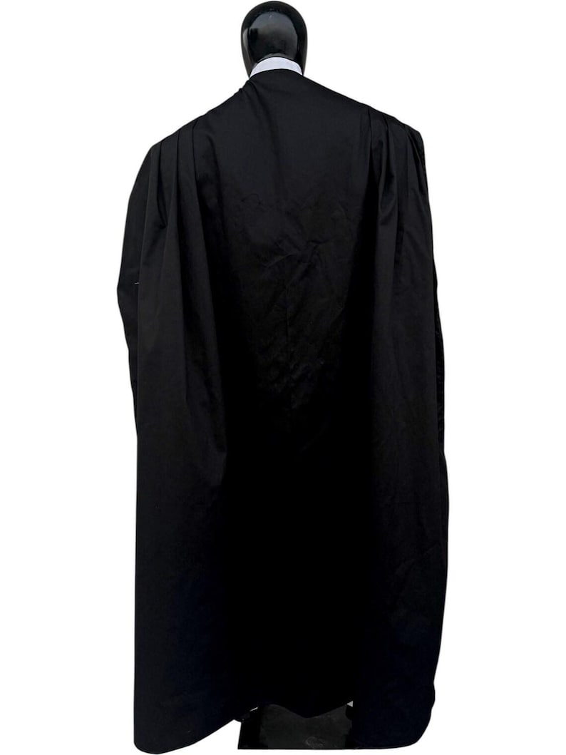 May include: A long, black robe with a high collar, displayed on a mannequin. The robe is made of a dark, opaque fabric and drapes down to the mannequin's feet. The mannequin's head is black and glossy, with a white collar visible.