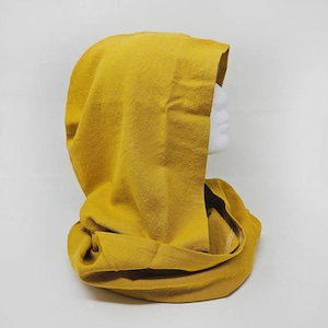 May include: Mustard yellow hood and cowl combination. The hood is draped over a white mannequin head. The cowl is wrapped around the neck. The fabric has a soft, textured appearance. The background is plain white.