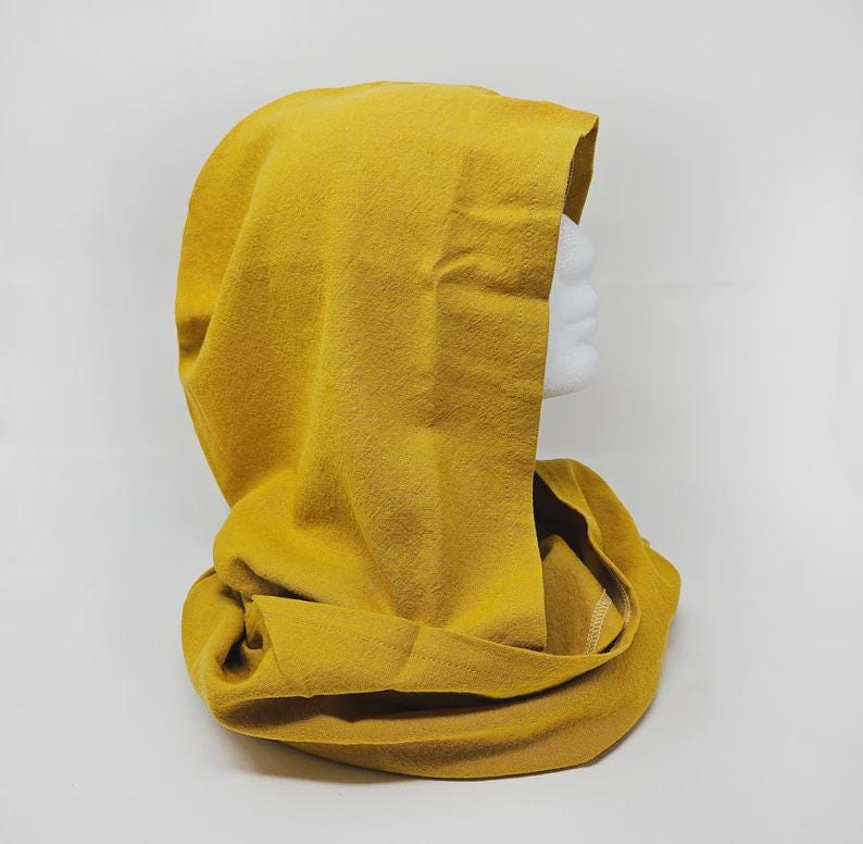 May include: Mustard yellow hood and cowl combination. The hood is draped over a white mannequin head. The cowl is wrapped around the neck. The fabric has a soft, textured appearance. The background is plain white.
