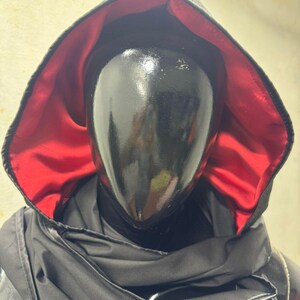 May include: A black hooded cloak with a red satin lining. The cloak is draped over a black mannequin head. The hood is up, and the cloak appears to be made of a shiny, possibly waterproof material. The overall aesthetic is dark and mysterious.
