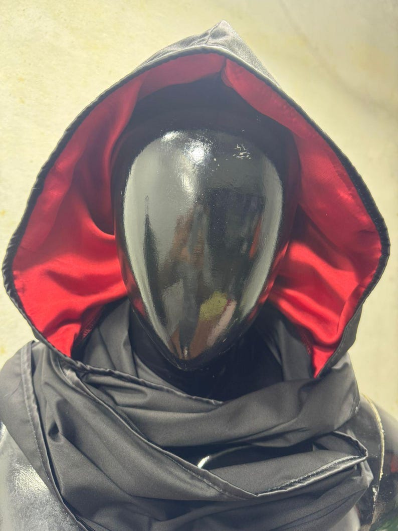 May include: A black hooded cloak with a red satin lining. The cloak is draped over a black mannequin head. The hood is up, and the cloak appears to be made of a shiny, possibly waterproof material. The overall aesthetic is dark and mysterious.