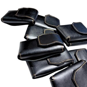 May include: A collection of black leather wallets with a flap closure and contrasting tan stitching. The wallets are stacked and arranged against a white background, showcasing their design and craftsmanship.