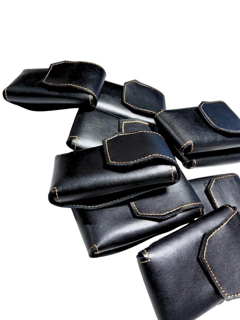May include: A collection of black leather wallets with a flap closure and contrasting tan stitching. The wallets are stacked and arranged against a white background, showcasing their design and craftsmanship.