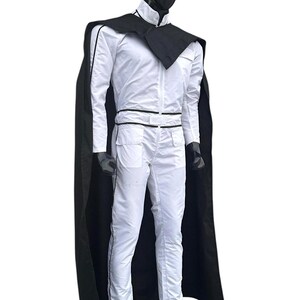 May include: A white jumpsuit with black trim and a matching black cape. The outfit features a high collar, a zip-up front, and pockets. The mannequin is wearing black gloves and shoes.