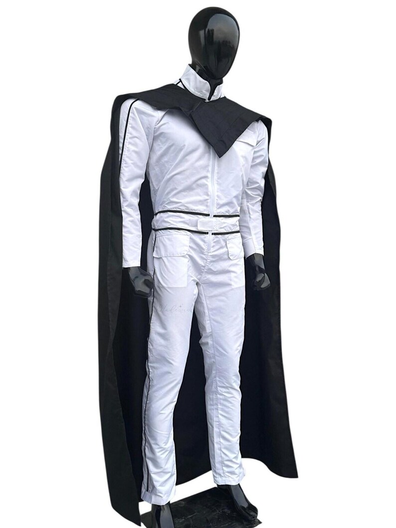 May include: A white jumpsuit with black trim and a matching black cape. The outfit features a high collar, a zip-up front, and pockets. The mannequin is wearing black gloves and shoes.