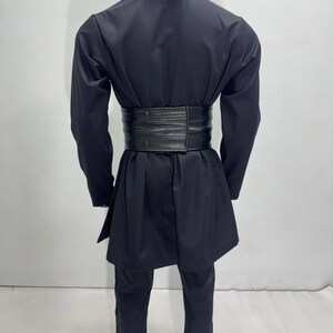 Op de afbeelding: Een zwarte outfit bestaande uit een jas met lange mouwen, een broek en een brede riem. De jas heeft een opstaande kraag en een wikkel sluiting. De brede riem accentueert de taille. De broek heeft rechte pijpen.