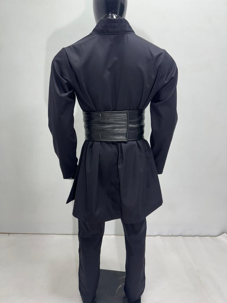 Op de afbeelding: Een zwarte outfit bestaande uit een jas met lange mouwen, een broek en een brede riem. De jas heeft een opstaande kraag en een wikkel sluiting. De brede riem accentueert de taille. De broek heeft rechte pijpen.