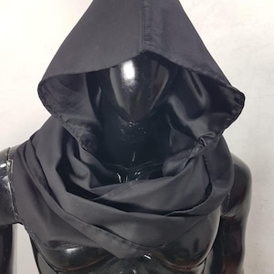 May include: A black hooded cowl made of a soft, draping fabric. The hood is pointed and the cowl wraps around the neck. The garment is displayed on a black mannequin, showcasing its shape and design. The overall aesthetic is dark and mysterious.