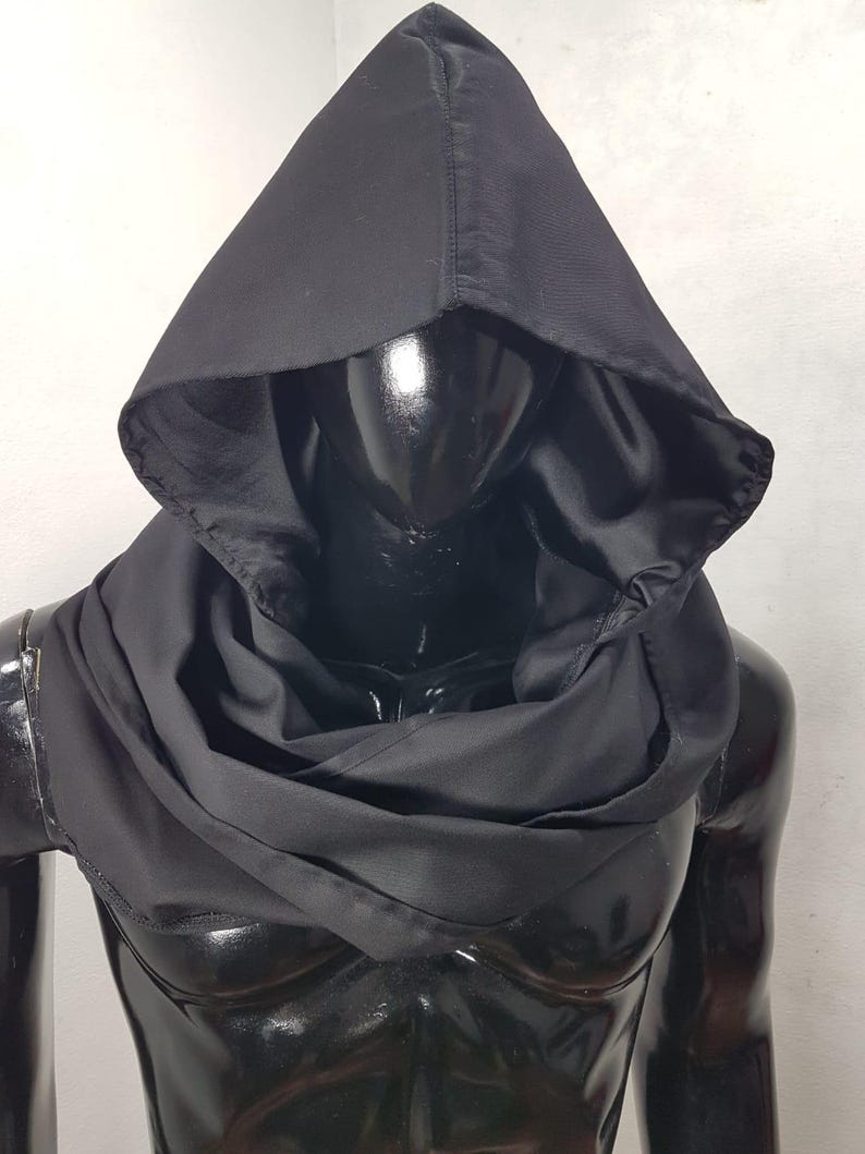 May include: A black hooded cowl made of a soft, draping fabric. The hood is pointed and the cowl wraps around the neck. The garment is displayed on a black mannequin, showcasing its shape and design. The overall aesthetic is dark and mysterious.