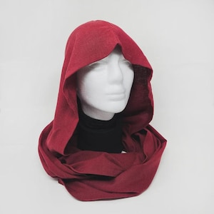 May include: A deep red hooded cowl, displayed on a white mannequin head. The cowl is made of a soft, textured fabric and features a hood and a long, scarf-like section that wraps around the neck. The mannequin is wearing a black turtleneck.