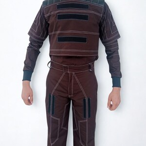 May include: A brown two-piece outfit with white stitching and black accents. The top has a high collar and short sleeves, with three black rectangular patches. The pants have matching white stitching and two black rectangular patches.