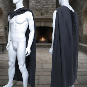 Puede incluir: Una capa gris oscuro drapeada sobre un maniqu&iacute;, mostrada de frente y de espaldas. La capa es larga, llegando a los tobillos del maniqu&iacute;. El fondo presenta una pared de piedra y una chimenea, lo que sugiere un tema medieval o de fantas&iacute;a.