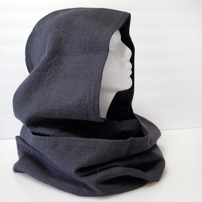 May include: A dark gray hooded cowl made of textured fabric. The cowl is draped over a white mannequin head, showcasing the hood's shape and the neck warmer design. The fabric appears to be linen or a similar material.