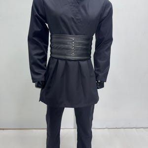 Op de afbeelding: Zwart kostuum met een wikkel-top, lange mouwen en bijpassende broek. Een brede, zwarte riem met decoratieve klinknagels accentueert de taille. De outfit wordt gepresenteerd op een zwarte mannequin, wat wijst op een cosplay- of theaterthema.