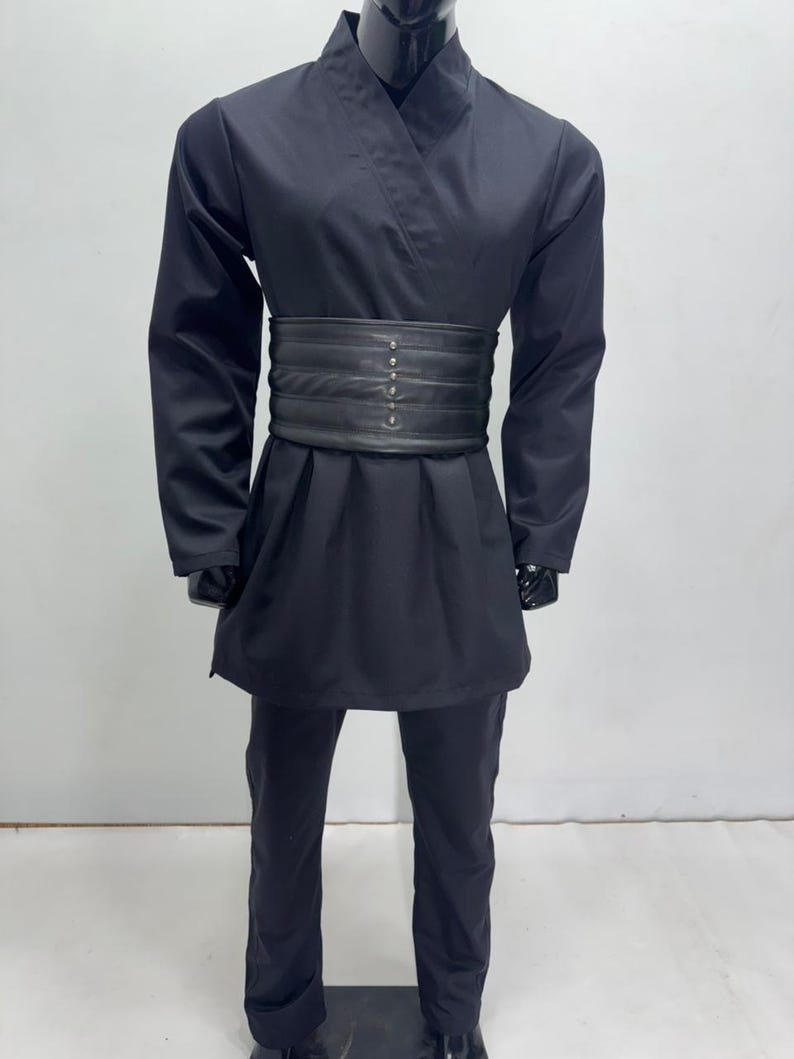 Op de afbeelding: Zwart kostuum met een wikkel-top, lange mouwen en bijpassende broek. Een brede, zwarte riem met decoratieve klinknagels accentueert de taille. De outfit wordt gepresenteerd op een zwarte mannequin, wat wijst op een cosplay- of theaterthema.