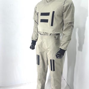 Puede incluir: Un traje futurista de color beige claro exhibido en un maniqu&iacute;. El atuendo incluye una parte superior de cuello alto y pantalones a juego, ambos con detalles rectangulares negros y costuras blancas. Guantes negros completan el look.