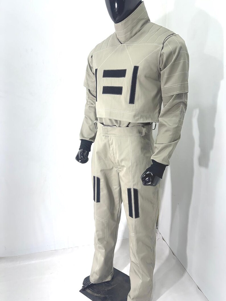 Puede incluir: Un traje futurista de color beige claro exhibido en un maniqu&iacute;. El atuendo incluye una parte superior de cuello alto y pantalones a juego, ambos con detalles rectangulares negros y costuras blancas. Guantes negros completan el look.