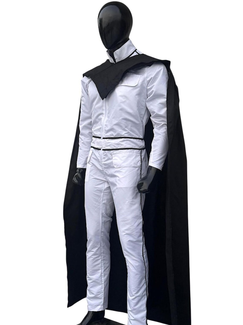 May include: A white jumpsuit with black trim and a matching black cape. The outfit is displayed on a mannequin with a black head. The jumpsuit has a high collar and a black belt. The cape drapes over the shoulders.