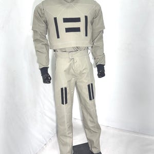 Puede incluir: Un traje futurista de color beige claro exhibido en un maniqu&iacute;. El atuendo incluye un top corto con acentos rectangulares negros, pantalones a juego y un cuello alto. Guantes y botas negras completan el look. Las costuras blancas delinean las costuras.