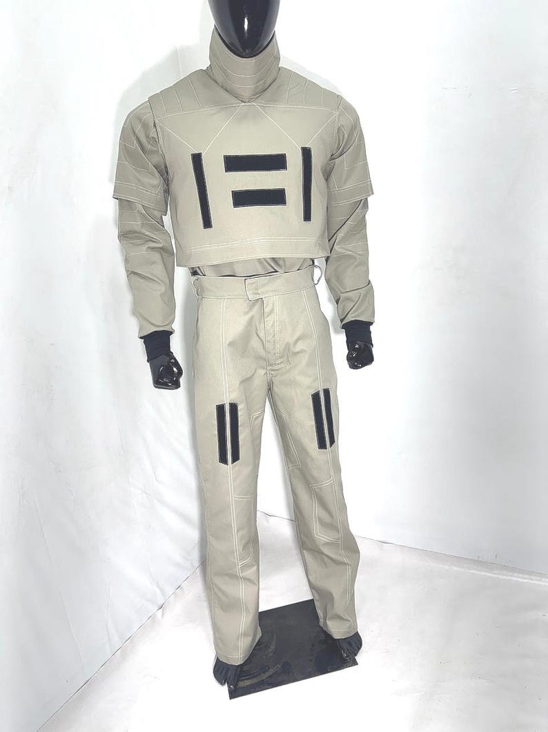 Puede incluir: Un traje futurista de color beige claro exhibido en un maniqu&iacute;. El atuendo incluye un top corto con acentos rectangulares negros, pantalones a juego y un cuello alto. Guantes y botas negras completan el look. Las costuras blancas delinean las costuras.