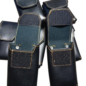 May include: Several black leather pouches with gold stitching are displayed. Two pouches are open, revealing a dark interior and a Velcro closure. The pouches have a rectangular shape with rounded edges and a flap closure.