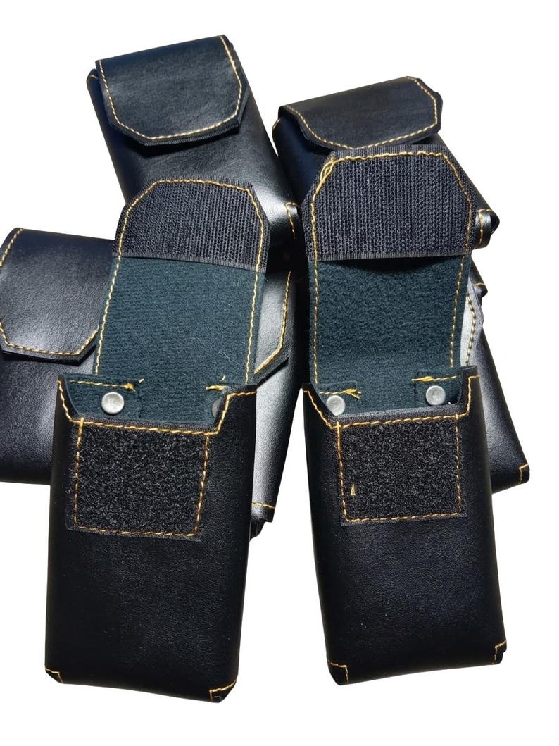 May include: Several black leather pouches with gold stitching are displayed. Two pouches are open, revealing a dark interior and a Velcro closure. The pouches have a rectangular shape with rounded edges and a flap closure.