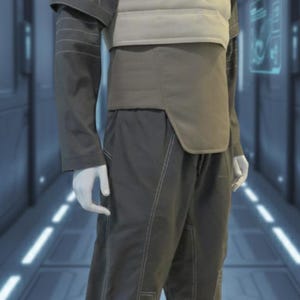 Puede incluir: Maniqu&iacute; con un atuendo gris futurista. El conjunto incluye una camisa de cuello alto, un chaleco acolchado, un cintur&oacute;n y pantalones a juego. Las costuras blancas acent&uacute;an las costuras. El atuendo se exhibe sobre una plataforma plateada.