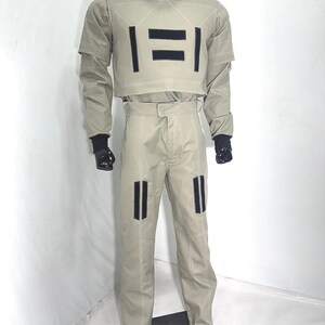 Puede incluir: Un disfraz color beige con detalles negros. El conjunto incluye una parte superior de cuello alto con detalles rectangulares negros y pantalones a juego. El disfraz tiene guantes negros y un cubrecabezas negro. Se ven costuras blancas.