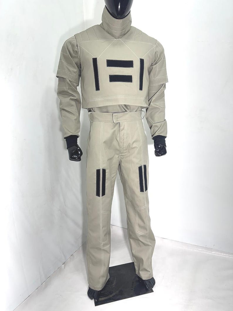 Puede incluir: Un disfraz color beige con detalles negros. El conjunto incluye una parte superior de cuello alto con detalles rectangulares negros y pantalones a juego. El disfraz tiene guantes negros y un cubrecabezas negro. Se ven costuras blancas.